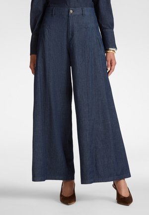 Frau trägt hoch taillierte, weit geschnittene dunkle Denim-Hosen mit vorderen Taschen und braunen spitzen Absätzen, steht vor einem schlichten Hintergrund.