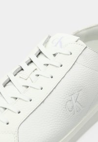 Close-up of white textured leather sneaker with white laces and subtle "CK" logo embossed on side and printed on tongue.