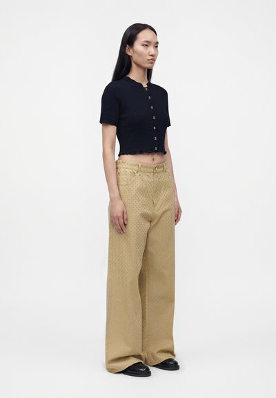 Γυναίκα στέκεται φορώντας μαύρο κοντό κουμπωτό πουκάμισο και μπεζ παντελόνια φαρδιά με μικρό πουά σχέδιο και μαύρα παπούτσια.