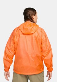 Orange, lätt regnjacka med huva, lös passform och elastiska ärmmuddar. Har en slät yta och en minimalistisk design.