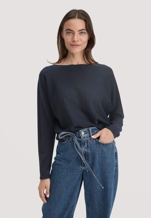 Frau mit schulterlangem braunem Haar trägt einen lockeren dunkelblauen Pullover und hoch taillierte blaue Jeans mit einem gebundenen Gürtel, die Hand in der Tasche.