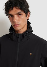 Czarna kurtka z wysokim kołnierzem, przednim zamkiem błyskawicznym i sznurkiem, z pomarańczowym logo na klatce piersiowej. Gładka tekstura materiału.