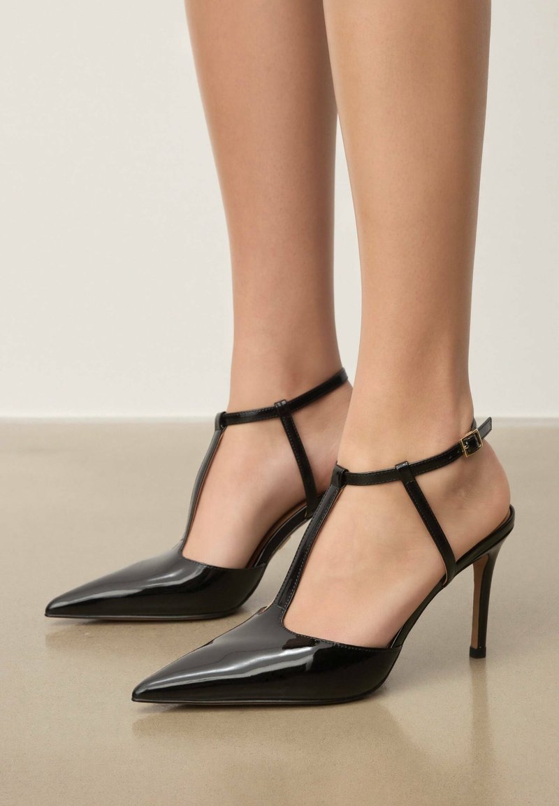 Escarpins noirs à bout pointu avec une finition en vernis brillant, dotés d'une bride en T et d'une boucle à la cheville, reposant sur un talon stiletto fin.