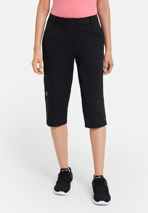 Frau trägt schwarze, knielange Freizeithose, ein pinkes Oberteil und schwarze Turnschuhe, steht vor weißem Hintergrund.