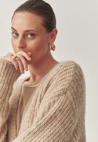 Beige, laza szabású kötött kardigán, puha tapintással. Arany fülbevalók absztrakt formával és hozzáillő arany gyűrű.