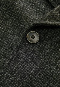 Tejido gris oscuro con un tejido texturizado, que presenta un botón negro redondo con un patrón en forma de cruz. Vista en primer plano del material y el detalle del cierre.