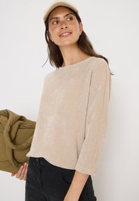 Top beige à côtes avec des manches trois quarts, col rond et texture douce. Le modèle tient un sac matelassé vert.