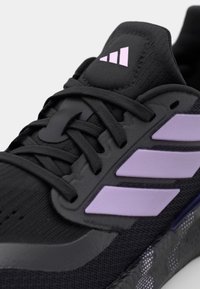 Chaussure de sport noire en tissu texturé, accents violets, lacets épais et trois bandes blanches sur le côté. Semelle incurvée.