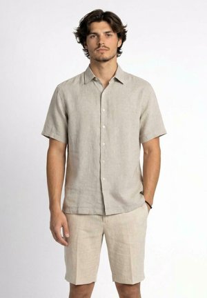 Giovane uomo in piedi che indossa una camicia beige a maniche corte con bottoni e pantaloncini coordinati fino al ginocchio con una mano in tasca.