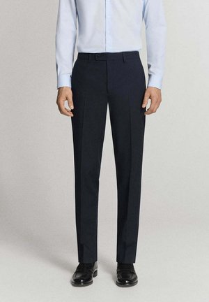 Uomo che indossa pantaloni eleganti neri, camicia azzurro chiaro a maniche lunghe e scarpe eleganti nere, in piedi contro uno sfondo neutro.