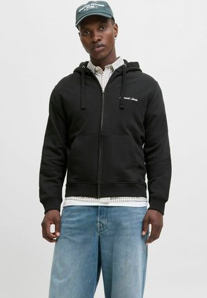 Sort zip-up hoodie med snoretræk, ribbede manchetter og kængurulomme, båret over en ternet skjorte og blå jeans.