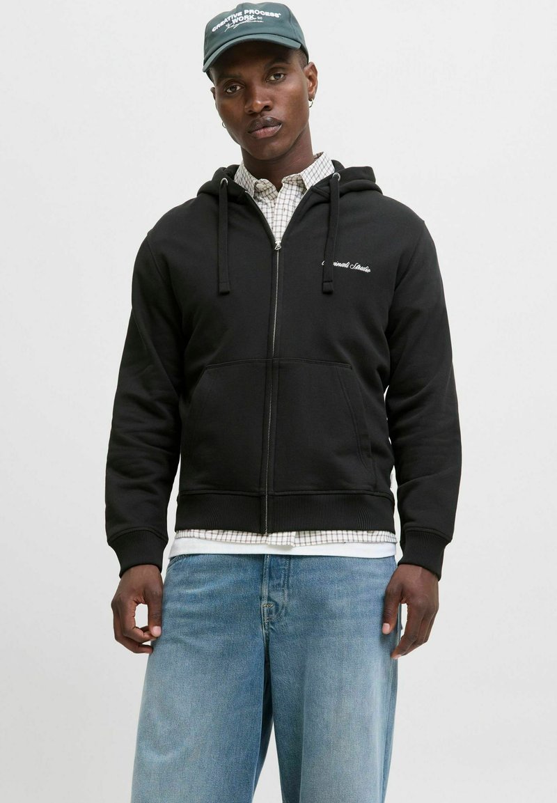 Sort zip-up hoodie med snoretræk, ribbede manchetter og kængurulomme, båret over en ternet skjorte og blå jeans.