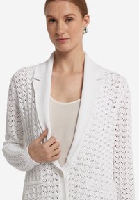 Weißer Strickcardigan mit einer strukturierten, offenen Webart, ausgestattet mit einem Schalkragen, einer durchgehenden Knopfleiste und Taschen, präsentiert vor einem neutralen Hintergrund.