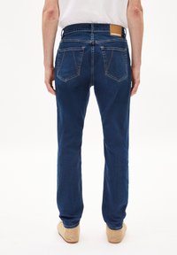Jeans de mezclilla azul oscuro con corte recto, diseño de cinco bolsillos y parche de cuero marrón en la cinturilla trasera. Textura suave.