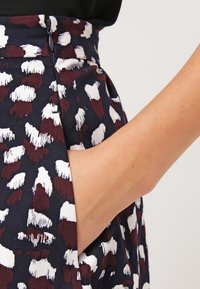 Sukně s vzorem, která má tmavě námořnické pozadí s bílými a burgundskými tvary připomínajícími štětcové tahy, s boční kapsou a detaily na zip.