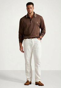 Moški nosi rjav dolgrokavni polo, bele hlače z pasom, rjave čevlje s pomponi in ure na zapestju, stoji ob preprosti ozadju.