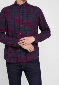 Tummanpunainen ja sininen ruudullinen pitkähihainen kauluksellinen nappipaita, jossa on yksittäinen rintatasku. Paidassa on pienet ruskeat napit.