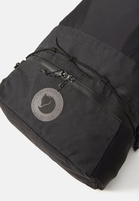 Zaino nero con una finitura texturizzata, tasca frontale con zip, chiusura con lacci e un patch circolare con logo. Materiale resistente con dettagli a contrasto.