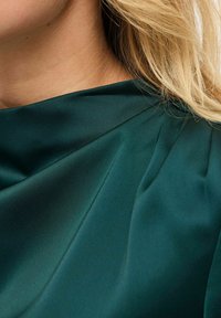 Dunkelgrüner Satinstoff drapiert sich elegant mit sanften Falten und zeigt eine glatte Textur sowie einen subtilen Glanz. Nahaufnahme des Halsbereichs.