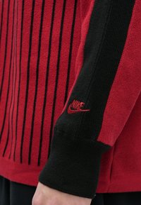 Piros és fekete pulóver függőleges fekete csíkokkal, bordázott textúrával, és a Nike logó piros színben a ujján. Normál szabás, hosszú ujjú.