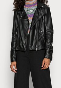 Schwarze Leder-Bikerjacke mit asymmetrischem Frontreißverschluss, schrägen Taschen und strukturierten Schulterakzenten. Getragen über einem bunten, gemusterten Oberteil.