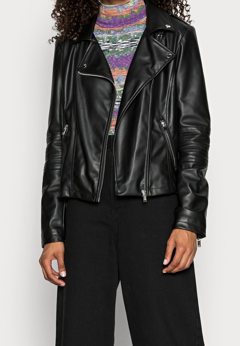 Schwarze Leder-Bikerjacke mit asymmetrischem Frontreißverschluss, schrägen Taschen und strukturierten Schulterakzenten. Getragen über einem bunten, gemusterten Oberteil.