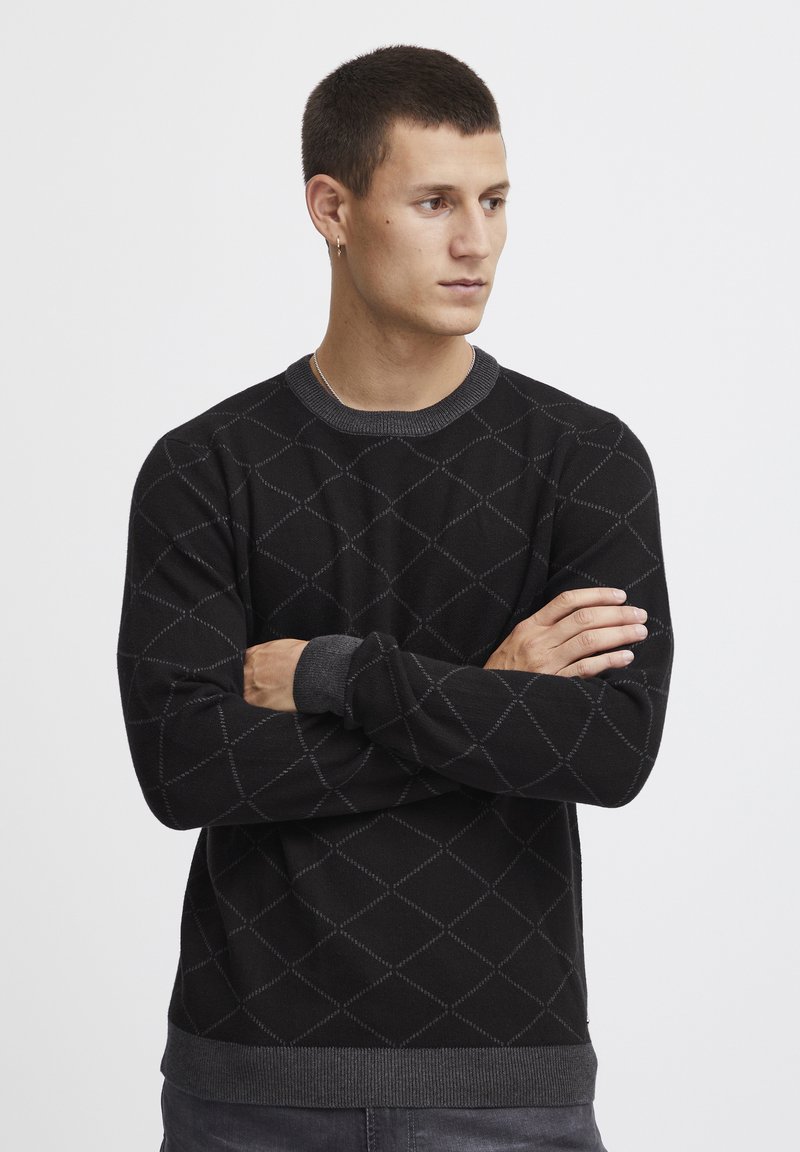 Schwarzer Pullover mit Rautenmuster, Rundhalsausschnitt und gerippten Bündchen. Das Material wirkt weich, mit kontrastierenden grauen Details am Kragen und Saum.