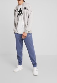 Sweatshirt zippé gris clair avec le logo "adidas", associé à un pantalon de jogging bleu arborant la marque "adidas" en blanc et des sneakers blanches.