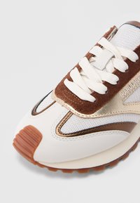 Witte sneaker met bruine suède en metallic accenten, witte veters en een gestructureerde rubberen zool, met het merk Karl Lagerfeld op de zijkant en de neuskap.