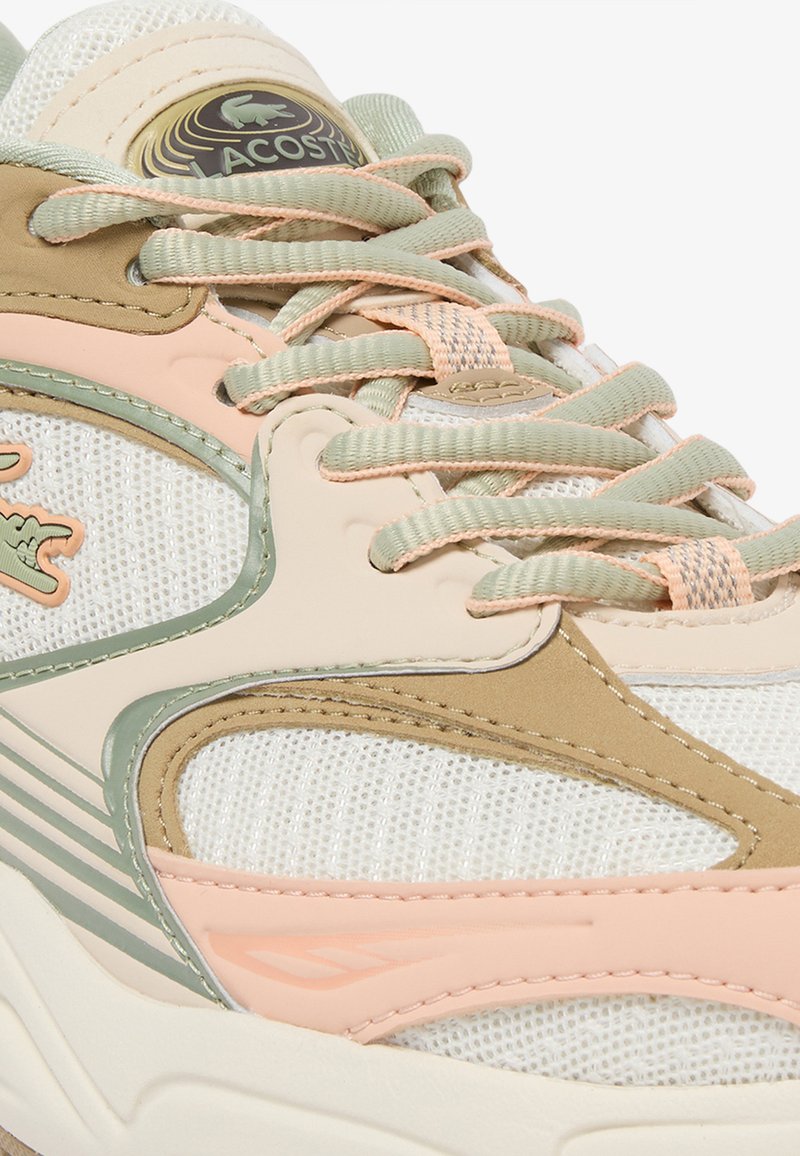 Nahaufnahme eines beigen, hellgrünen und pinken Lacoste-Sneakers mit Meshstoff, breiten Schnürsenkeln und sichtbarem Markenlogo auf der Zunge und der Seite.
