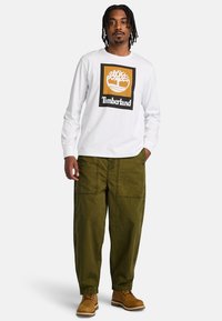 White long-sleeve shirt with black and orange Timberland logo, paired with olive green baggy pants and tan boots.