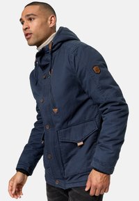 Marineblauwe jas met een capuchon, voorzien van een knoopsluiting, grote voorkantzak en leren accenten op de knopen en schouders.