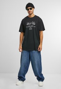 Black oversized t-shirt with white graphic text, paired with loose blue denim trousers and white sneakers, and a black knit beanie.