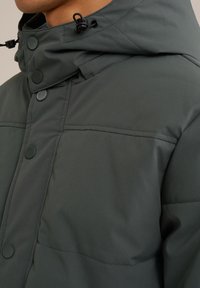 Närbild av en person som bär en mörkgrön vadderad jacka med huva, som visar knappar och sömnadsdetaljer på tyget.