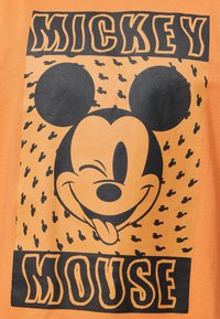 Oranžové tričko s velkým černým grafickým motivem Mickeyho Mouse, usmívajícího se s vyplazeným jazykem; na pozadí jsou malé černé siluety Mickeyho.