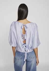 Bluză de un violet deschis cu mâneci bufante, trei șireturi la spate și un design cu spatele descoperit. Textură netedă cu un luciu subtil. Asortată cu blugi albaștri.