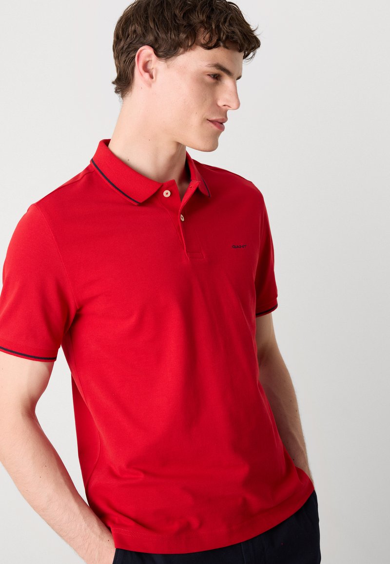 Polo rouge avec col, patte de boutonnage à trois boutons et accents bleu marine sur les poignets et le col. Présente un petit logo brodé sur la poitrine.