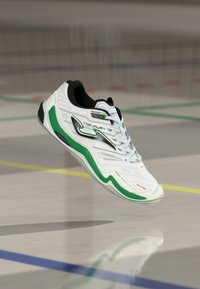 Scarpa sportiva bianca con accenti neri e verdi, caratterizzata da una superficie testurizzata, design di supporto e una suola ammortizzata per la stabilità.