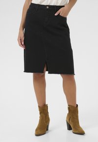 Jupe en denim noir avec une fente frontale et un ourlet effiloché. Le design comprend des passants de ceinture, des poches et une fermeture à bouton. Associée à des bottes à cheville marron.