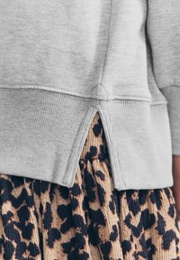 Pilkas džemperis su ribota apačia ir plyšiu nugaros pusėje, dėvimas ant sluoksniuoto sijono su smėlio spalvos ir juodo leopardžio raštu.