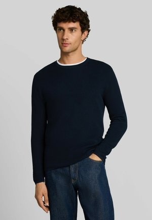 Man met krullend donker haar die een marineblauwe trui met lange mouwen draagt over een wit hemd en een blauwe spijkerbroek, met één hand in de zak.
