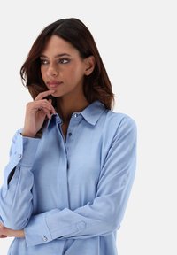 Hellblaues Button-up-Hemd mit Kragen, langen Ärmeln und sichtbaren Knöpfen. Der Stoff wirkt glatt und weich. Einfaches Design ohne Muster.