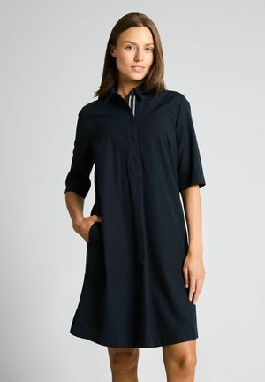 Jeune femme portant une robe-chemise noire ample mi-longue aux manches trois-quarts, debout devant un fond clair uni.