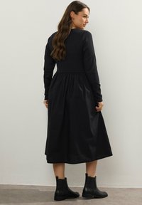 Nainen, jolla on pitkä hius, pukeutuneena mustaan pitkähihaiseen mekkoon ja mustiin nilkkabootseihin, seisoo ja katsoo olkansa yli tasaista seinää vasten.