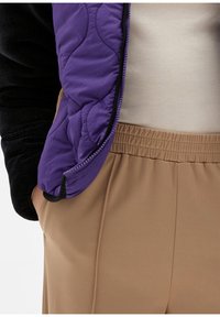 Paarse gewatteerde jas met zwarte fleece mouwen, gedragen over een lichtbeige top en beige elastische tailleband broeken. Naadloze stoftextuur zichtbaar.