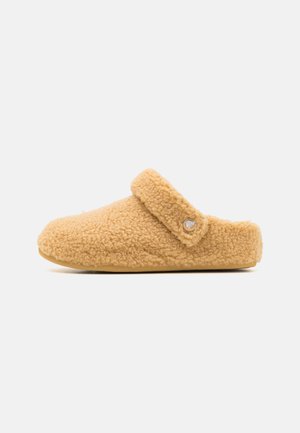 Fuzzy beige slipper with a soft texture, rounded shape, and a strap across the top. Features a small logo button on the side.