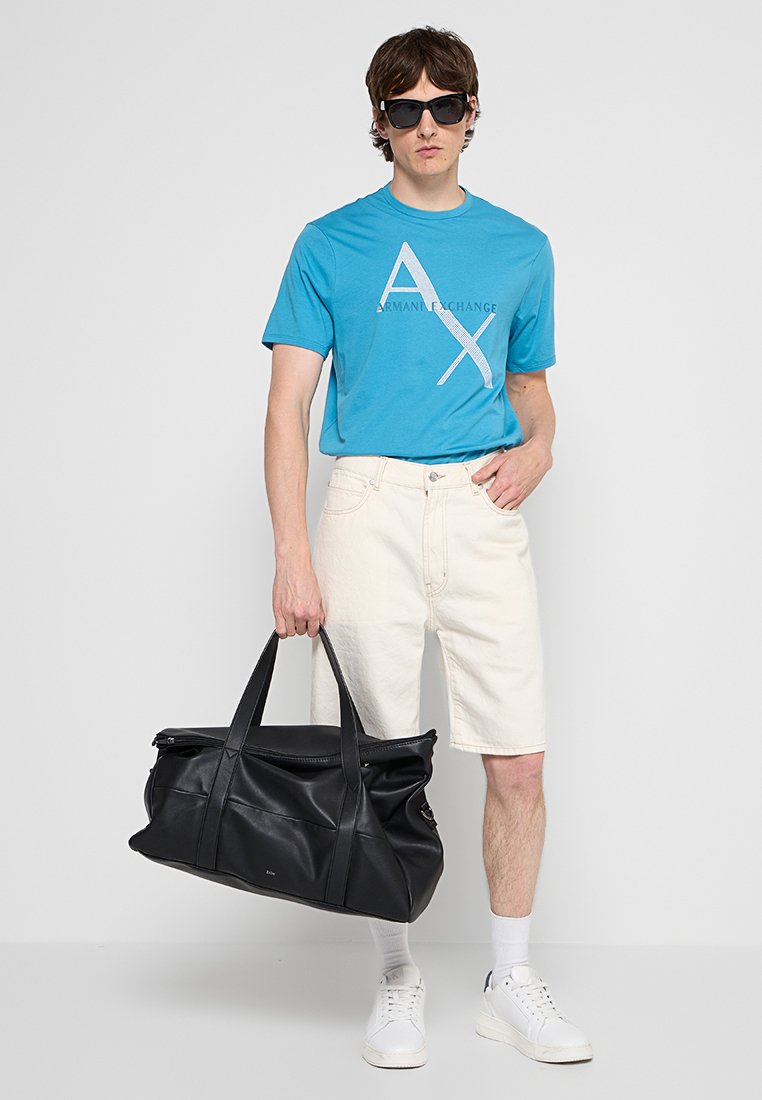 Uomo con occhiali da sole, t-shirt blu Armani Exchange, pantaloni corti bianchi, scarpe da ginnastica bianche, che tiene un grande borsone in pelle nera al lato.