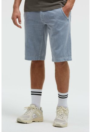 Mann trägt hellblaue Jeansshorts, weiße Socken mit schwarzen Streifen und beige Sneakers, steht mit einer Hand in der Tasche.