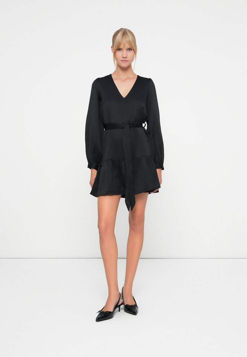 Vestido negro de tela ligera, con escote en V, mangas largas abullonadas, cintura ajustada con un lazo y falda en capas.