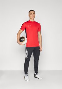 Rotes Nike-Sportshirt mit schwarzen Akzenten, schwarze NikeFC-Hosen mit weißem Logo und weiße Fußballschuhe, hält einen schwarz-weißen Fußball.